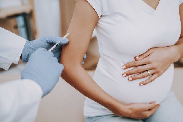 Eine schwangere Frau in einem weißen Hemd sitzt und hält ihren Bauch, während eine medizinische Fachkraft mit Handschuhen ihr eine Injektion in den Oberarm verabreicht.